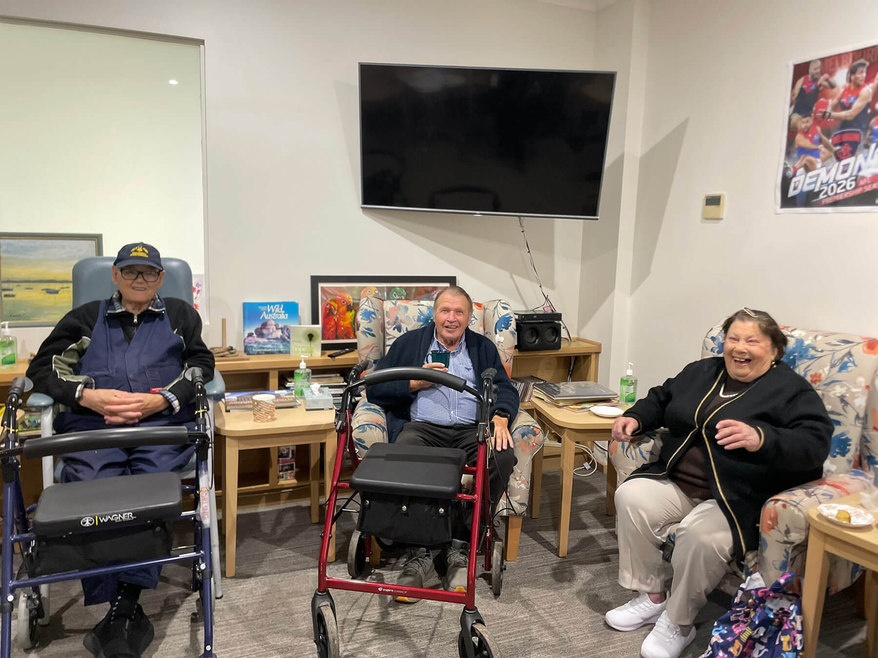 Three people sitting in armchairs in a day respite lounge-room. Two people have wheelie walkers infront of them.