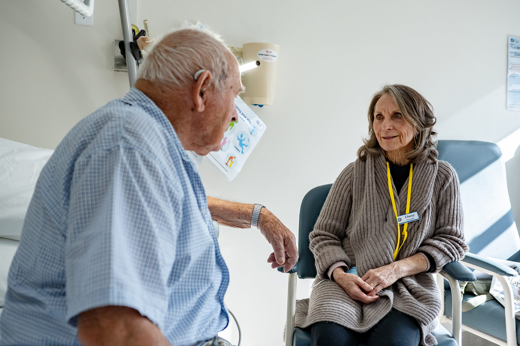 An elderly male patient sittingon the edge of his hospital bed, talking with a female volunteer at the hospital.